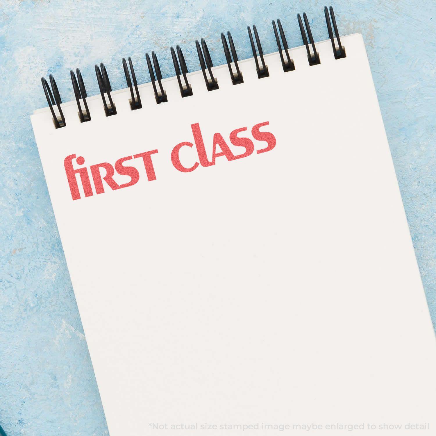 Bold Red First Class Xstamper Stamp imprinting First Class in red ink on a white spiral notebook against a blue background.