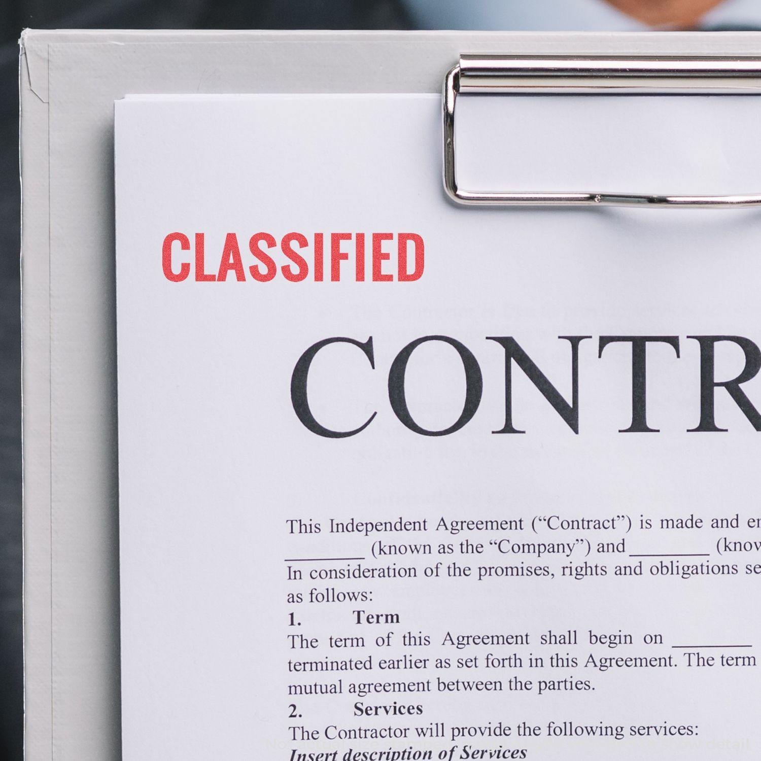 A document stamped with CLASSIFIED using a Classified Xstamper Stamp, held on a clipboard with visible contract text.