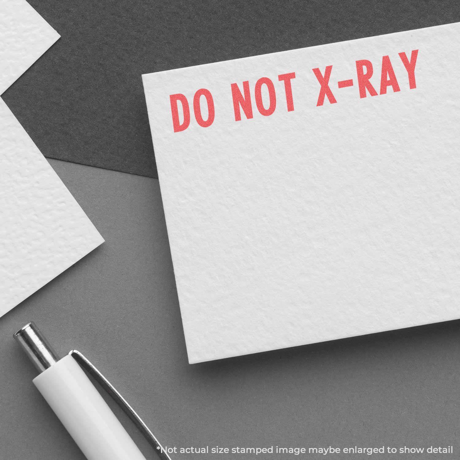 A white card stamped with DO NOT X-RAY in red ink using the Do Not X-Ray Xstamper Stamp, placed on a gray surface next to a pen.