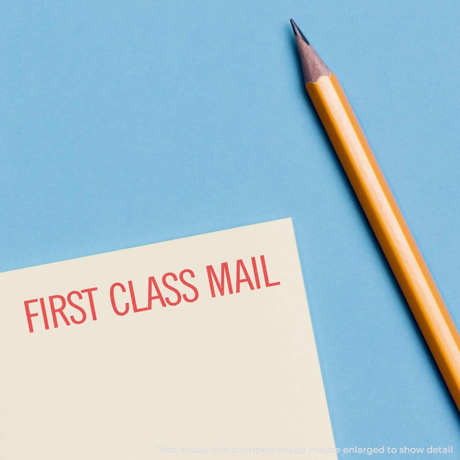 Jumbo Large Red First Class Mail Xstamper Stamp impression on a yellow paper next to a sharpened pencil on a blue background.