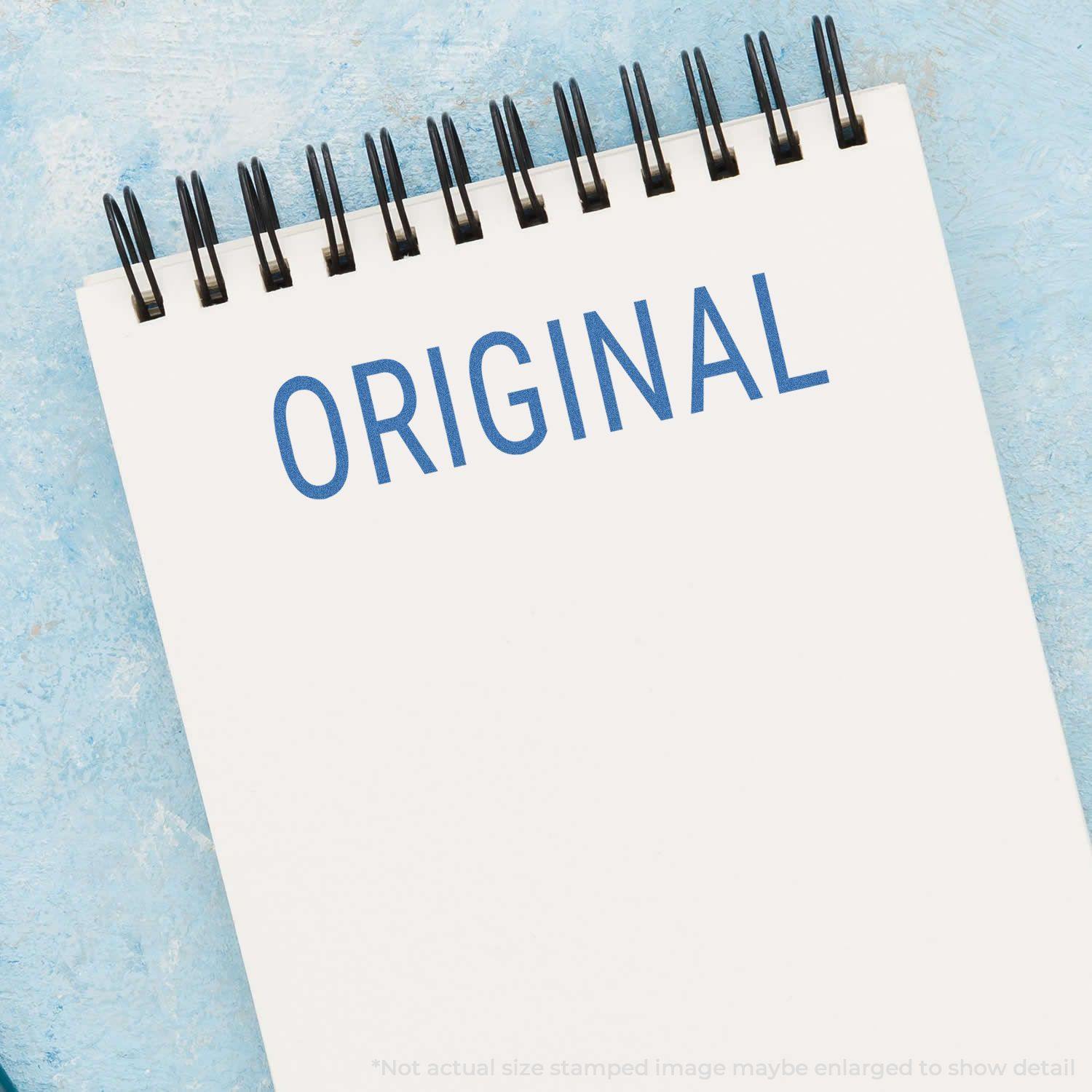 A spiral notebook with the word ORIGINAL stamped in blue using the Original Xstamper Stamp on a light blue textured background.