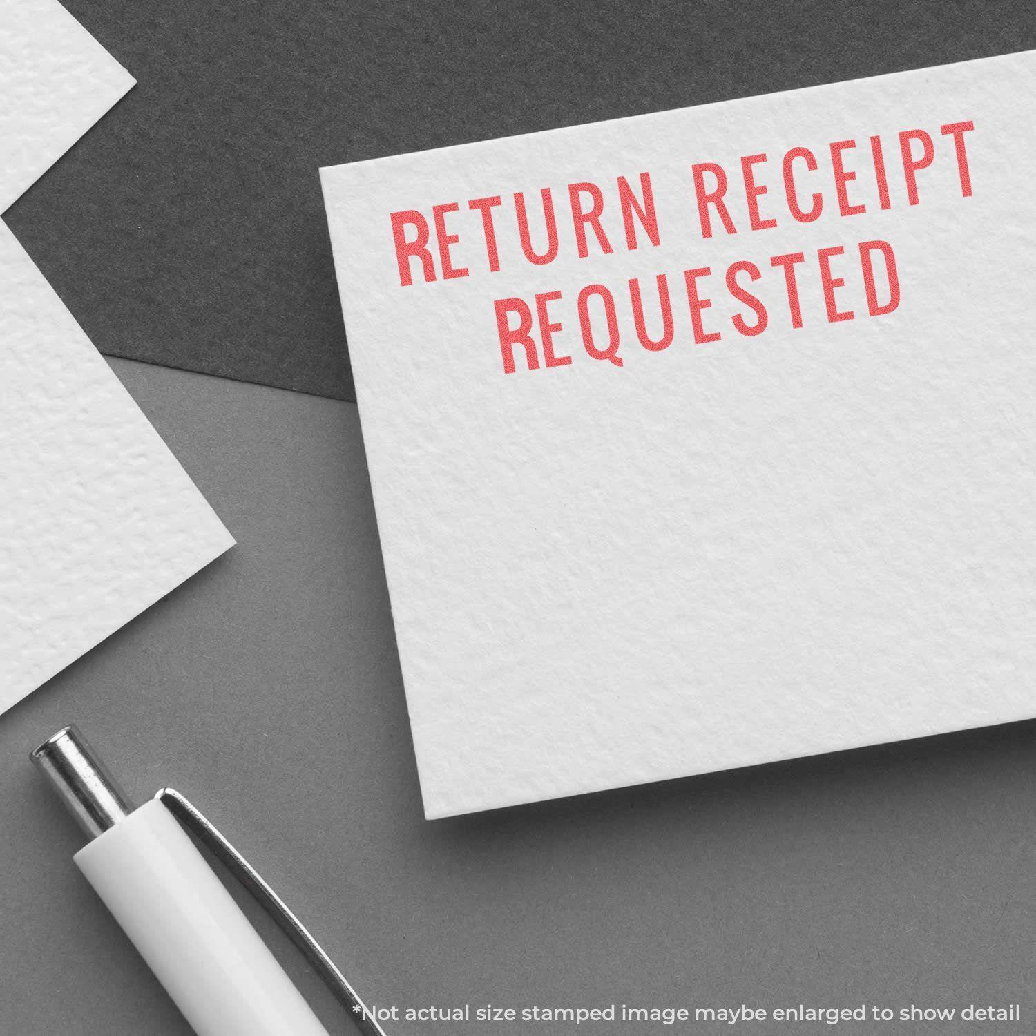 Return Receipt Requested Xstamper Stamp used on a white card, with a pen nearby on a gray surface.